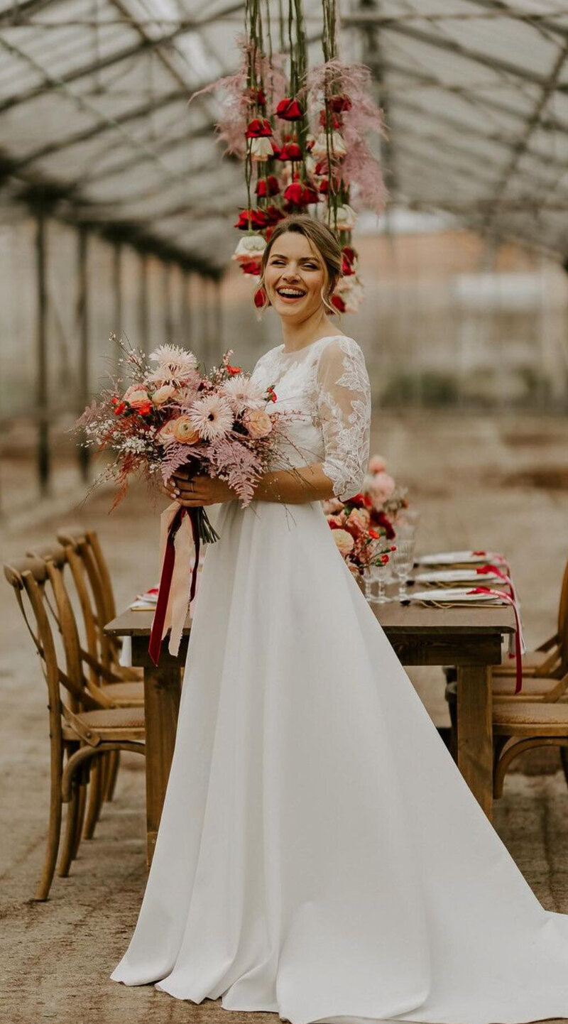 Mariée posant avec son bouquet devant une table en bois décorée et des fleurs suspendus à un plafond de verre pour un workshop photographie de mariage.