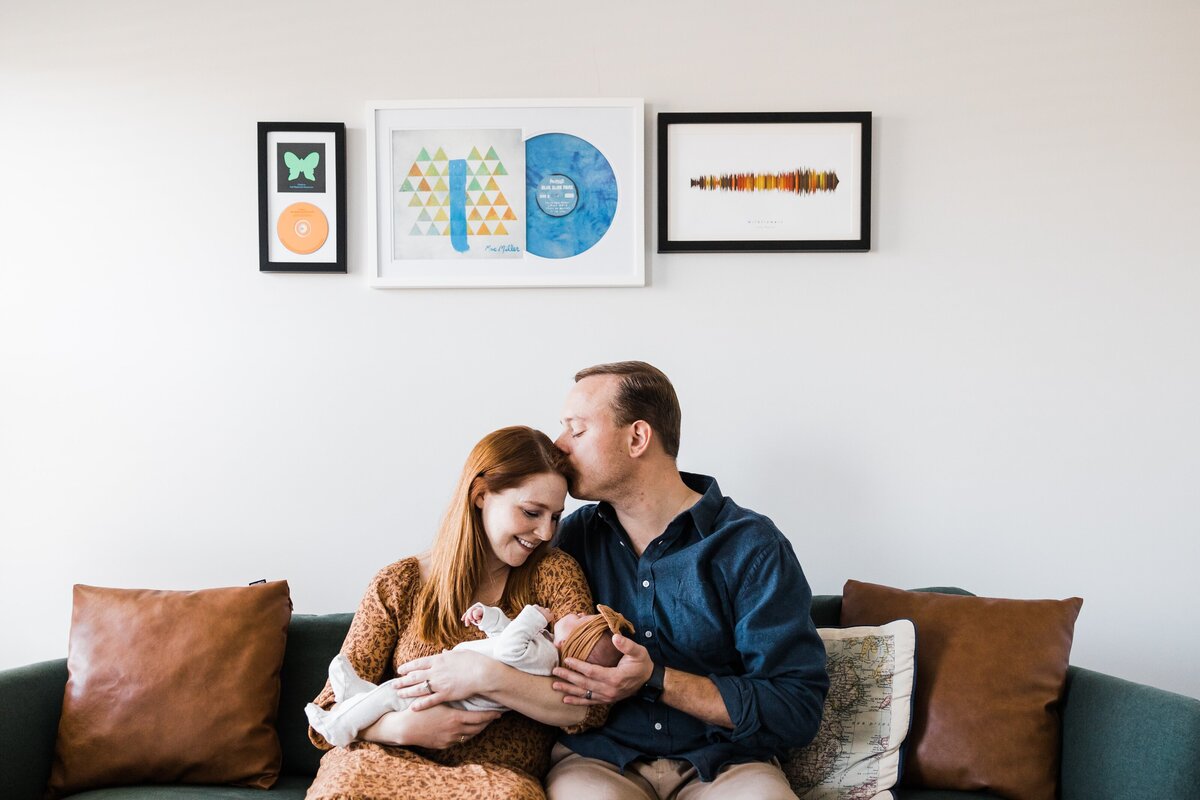 A couple sitting on a couch, affectionately embracing and gazing at a baby in their arms during an in-home newborn photography session, with colorful artwork hanging on the wall above them.