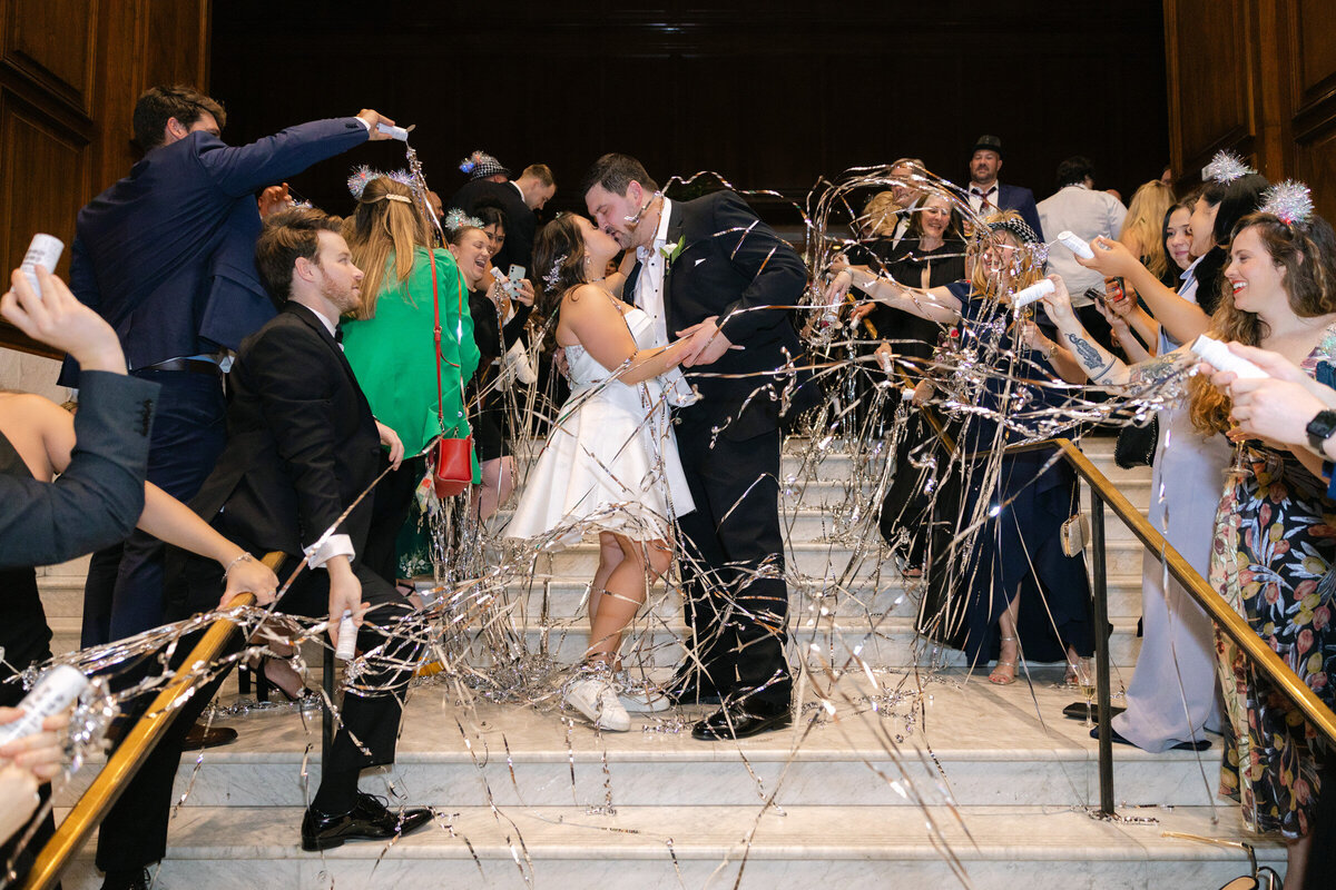 Bride and groom's exit from the wedding reception on the stair case bride and groom during their last private dance elegantly styled wedding reception table at The Adolphus in Dallas featuring a black tablecloth, tall glass vases with pink rose arrangements, numerous candles, and refined glassware and chargers
