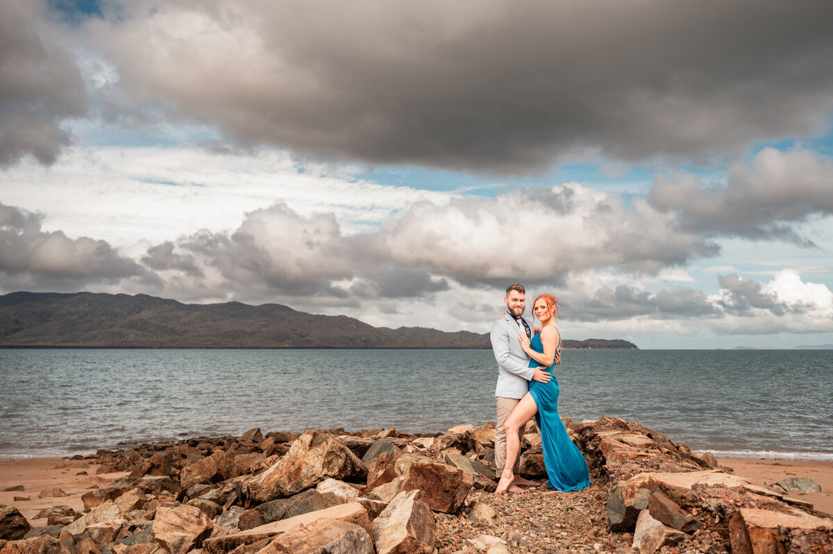 man in suit and woman in fancy blue dress embracing on a rock wall on a Townsville beach - Townsville Engagement Photography by Jamie Simmons