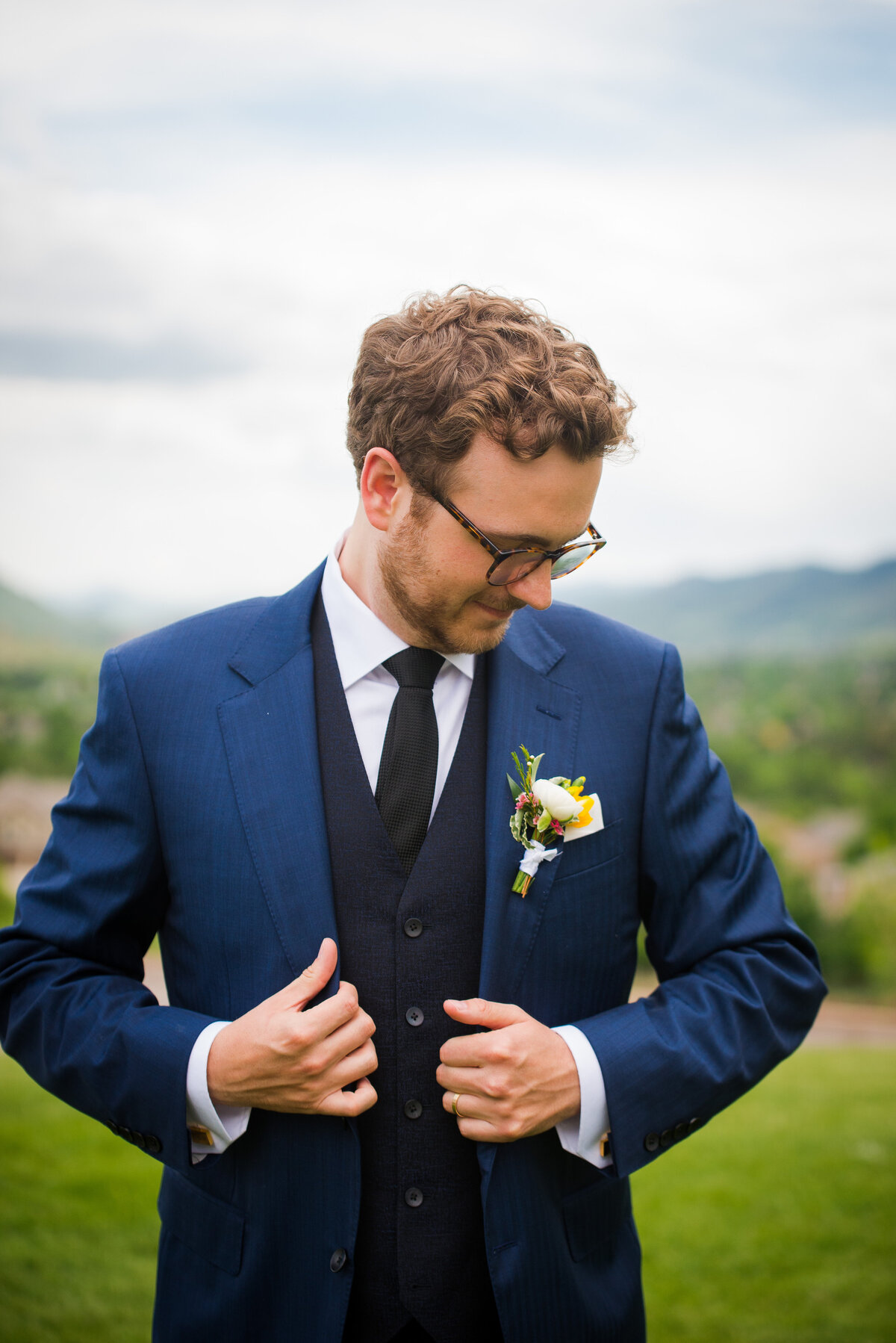 A groom adjusts his suit jacket and glances down at his boutonnière.