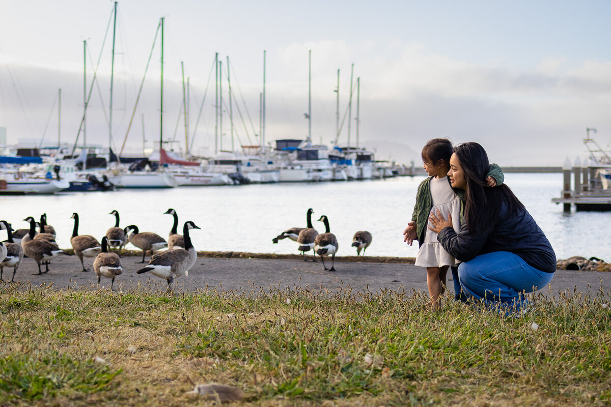 Mother and daughter crouch together by the water’s edge, watching geese at the harbor during a calm morning. Bay Area Family Portfolio - Ellobelle Photography