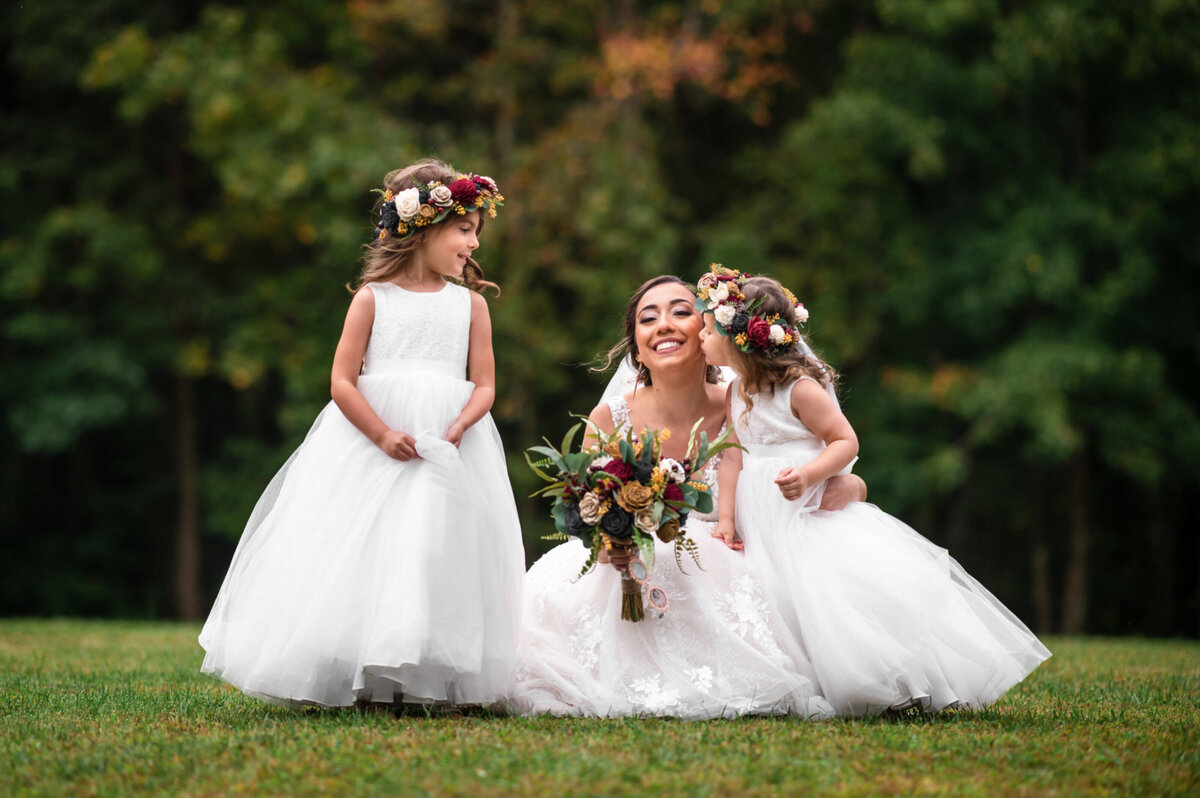 Flower girls wearing flower crowns kissing a bride on the cheek while taking bridal party photos at Brigalias