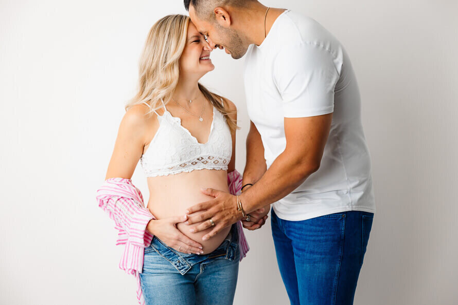 A man and his pregnant wife touch foreheads and put hand on her belly in the photo studio.