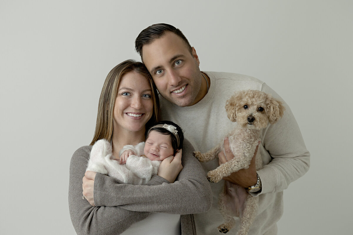 Happy new parents stand in a studio holding their smiling newborn daughter and small puppy