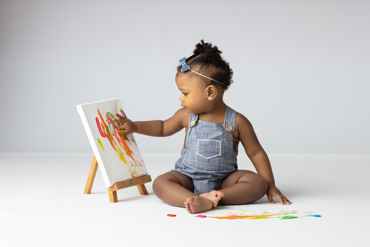 A toddler in denim overalls finger-paints on a small canvas, surrounded by splashes of colorful paint during a Dallas milestone photographer session.