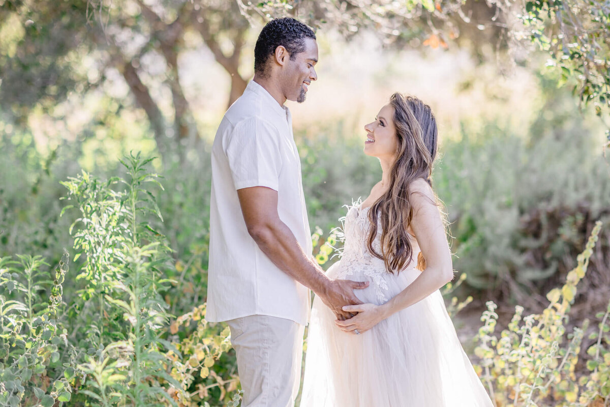 A couple stands together in a lush, sunlit garden. The man gently holds the womans pregnant belly as they gaze into each others eyes, both smiling softly. She wears a flowing white dress, while he is dressed in a white shirt and light pants.