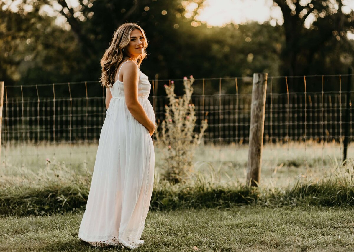 A pregnant woman in a white dress standing in a field at sunset captured by a Pittsburgh maternity photographer.