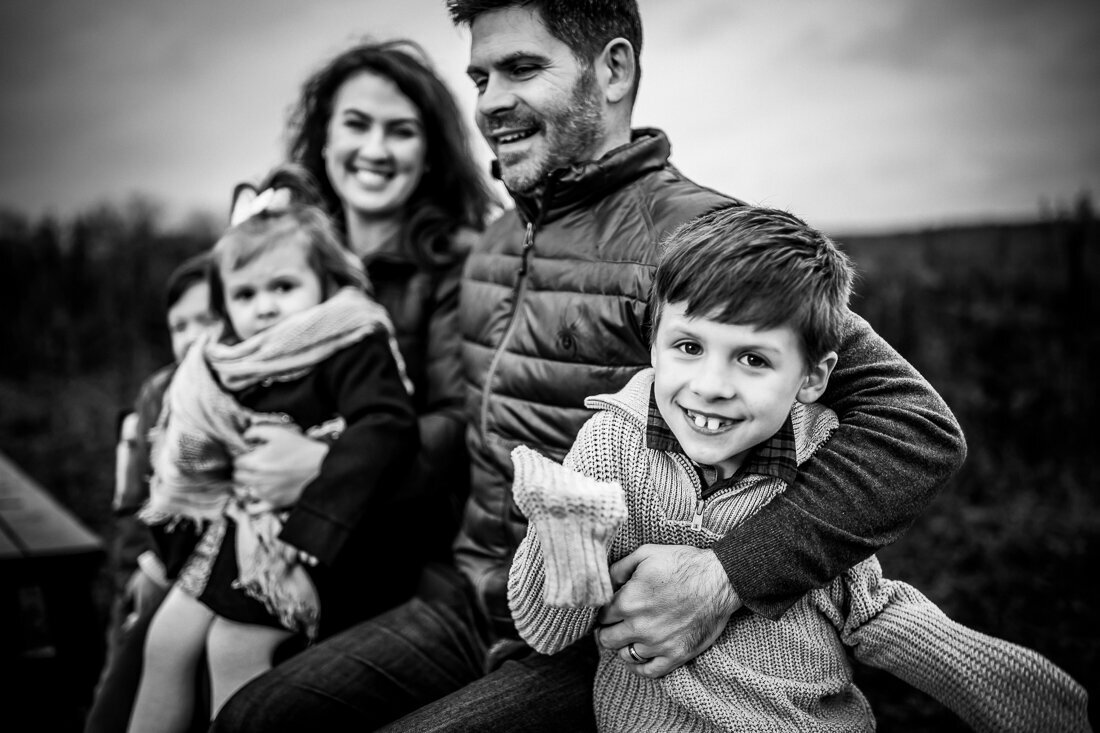 A black and white portrait of a family of four. Parents stand close together with the father holding a young boy and the mother holding a smaller child. The image captures them in an intimate, casual embrace outdoors.