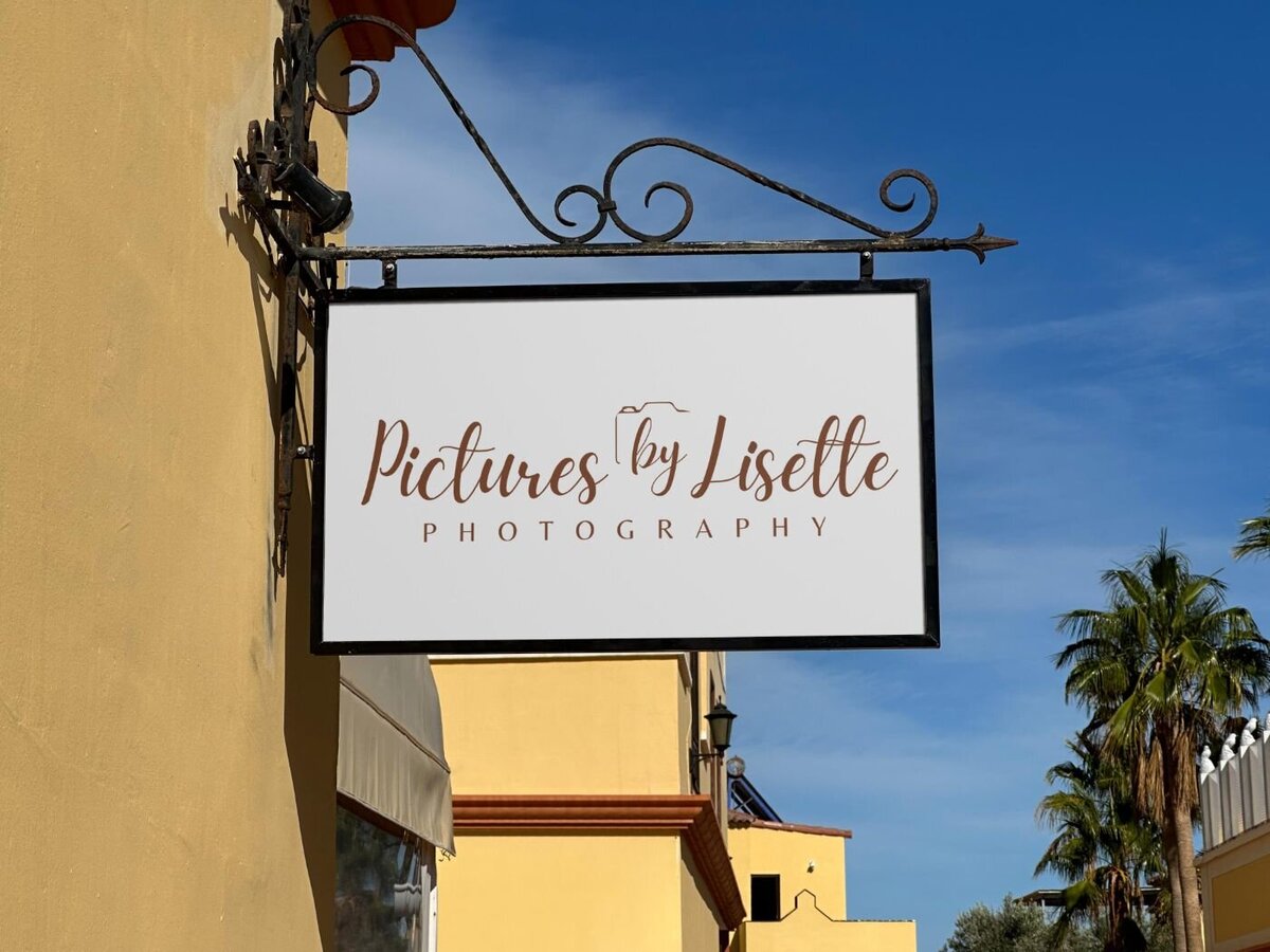 Outdoor metal hanging sign with ornate black bracket showing the logo “Pictures by Lisette Photography” in elegant brown script against a white background, set against a yellow building and blue sky.