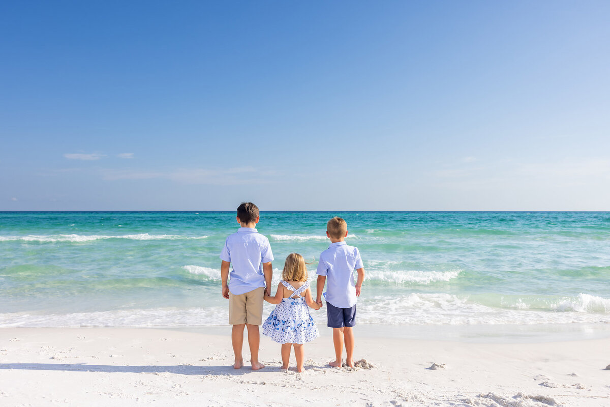 3 young siblings holding hands on the beach standing with their backs to the camera and looking out at the Gulf of America in Seagrove Beach, Florida.