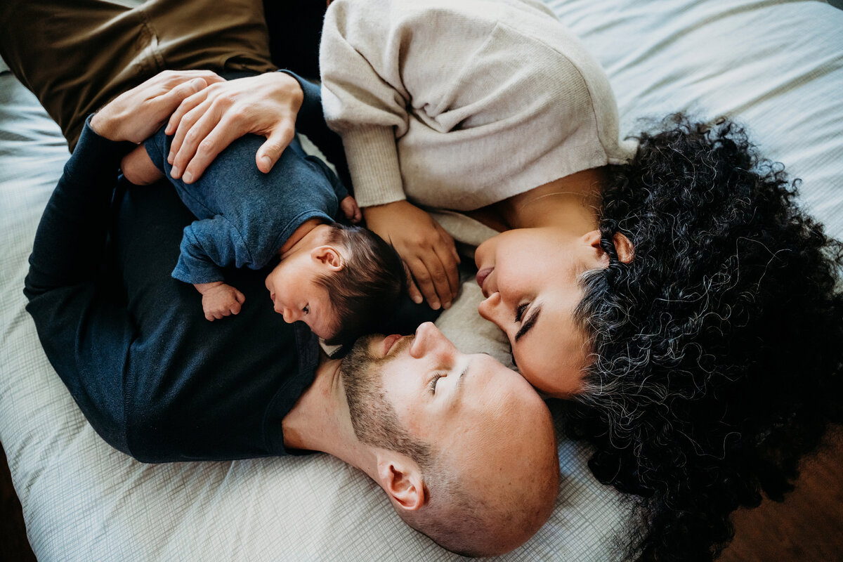 Newborn Photographer, husband and wife lean into each other tenderly as they lay on the bed holding baby