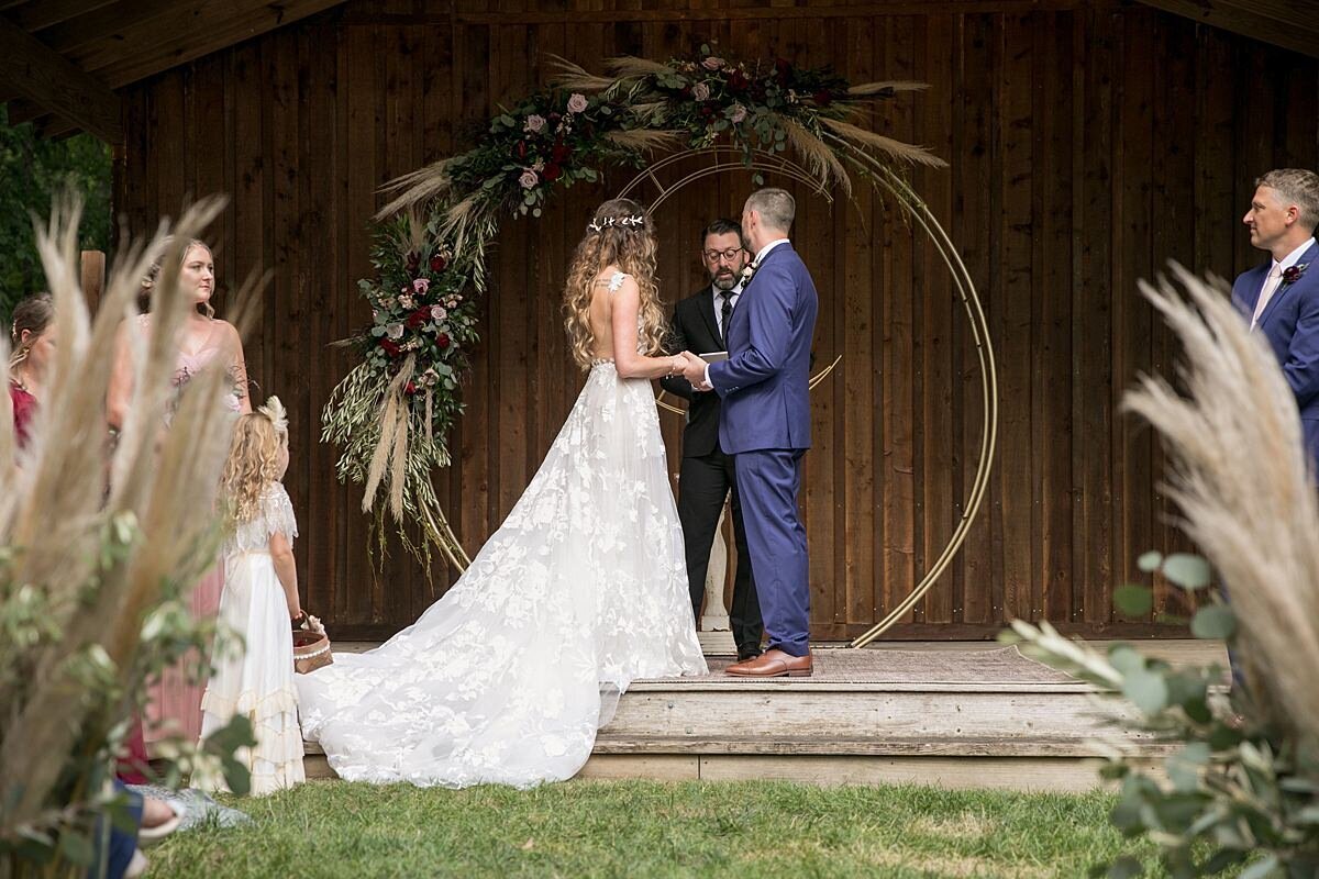 The bride, wearing a long lace wedding gown holds the groom's hands as they recite their wedding vows at the rustic log ceremony pavilion at Saddle Woods Farm. The groom is wearing a navy suit with a burgundy tie and the gold circular arbor is decorated with pampas grass, blush and burgundy flowers and greenery.