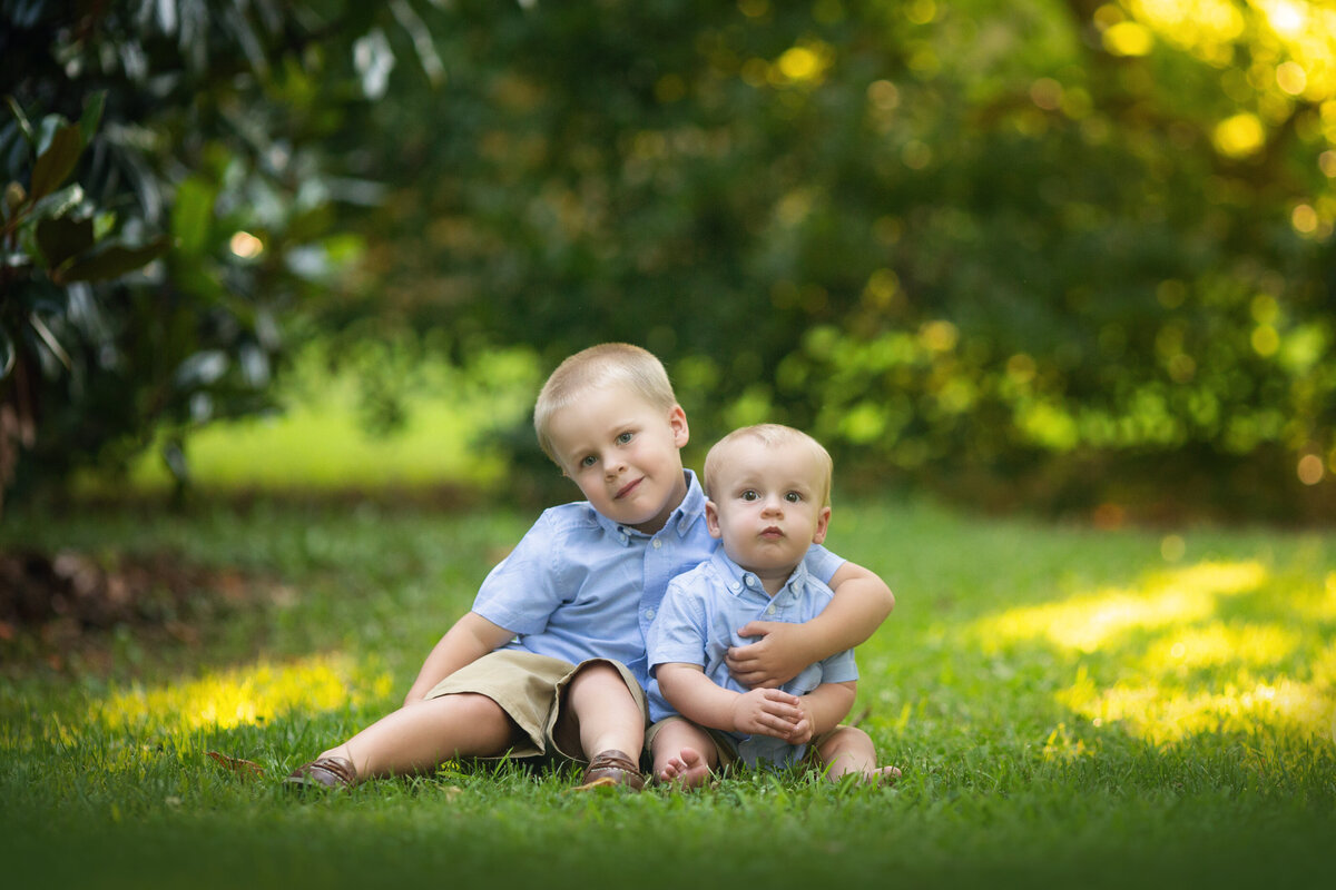 A young toddler boy hugs onto his baby brother in a matching shirt while sitting in a park lawn