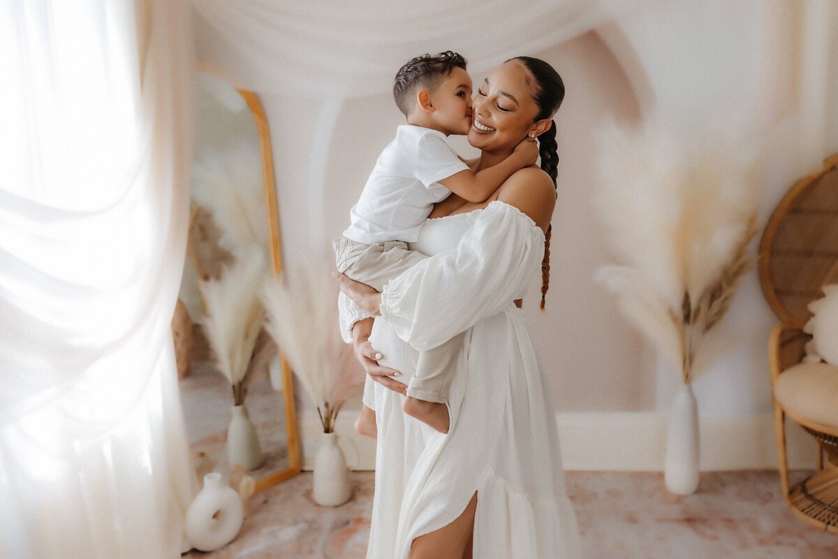 A woman in a white dress holds a smiling child in a softly lit room with pampas grass and draped fabric.