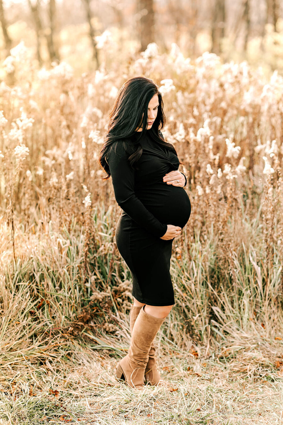 A pregnant woman in a fitted black dress and tan boots stands in a grassy field, cradling her belly amid golden plants under soft light—an elegant moment captured by a Bloomington maternity photographer specializing in pregnancy photography.