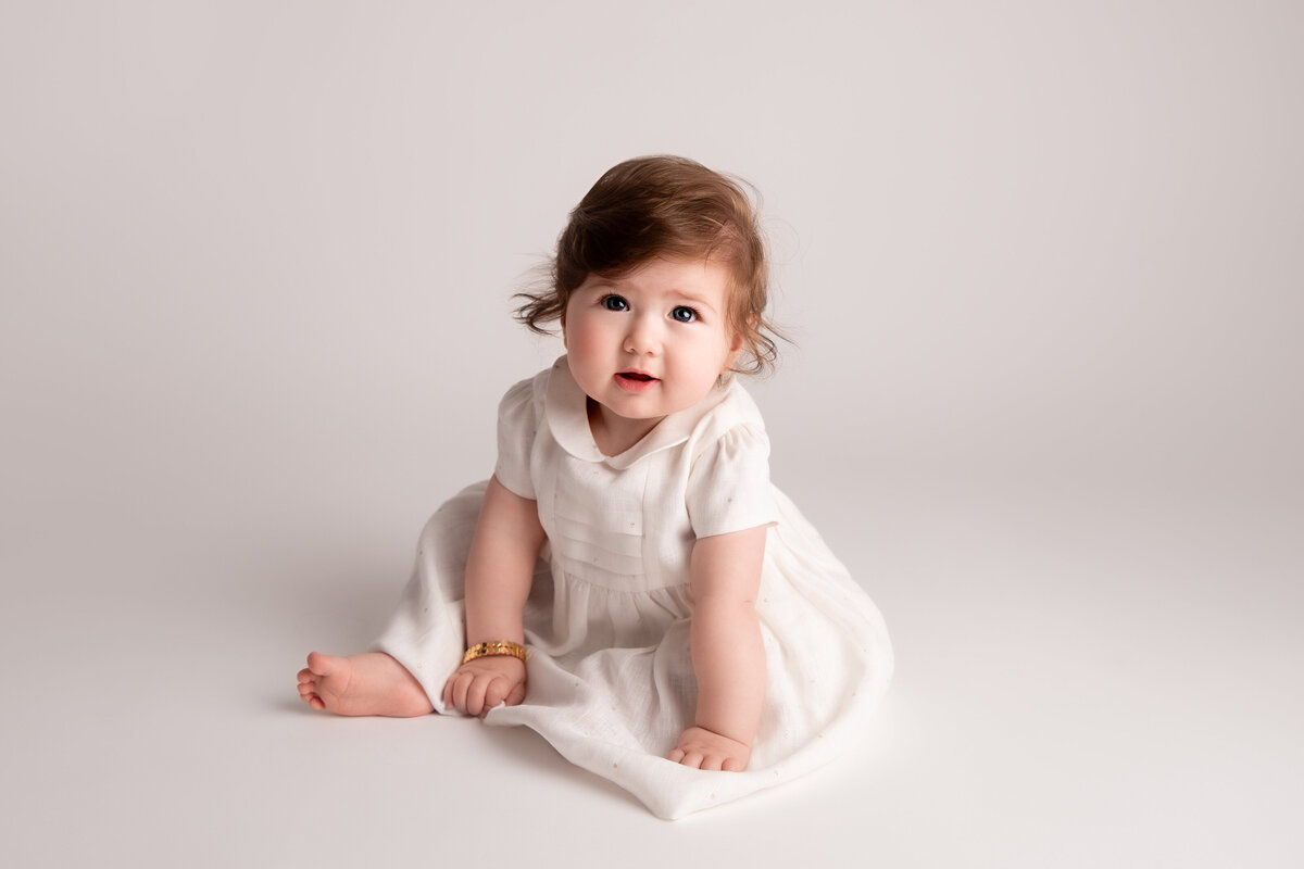 Six month old baby girl dressed in a white dress and leaning forward toward the camera smiling.