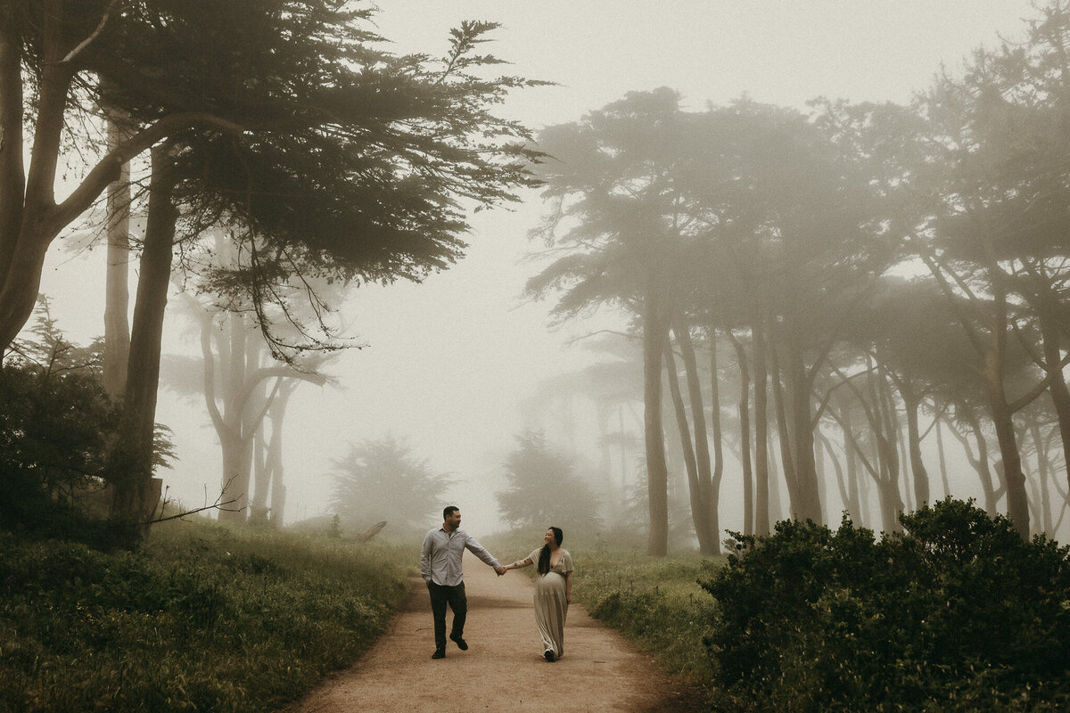 San Francisco maternity couple holds hands while walking along a tree-lined trail and Lands End on a foggy day