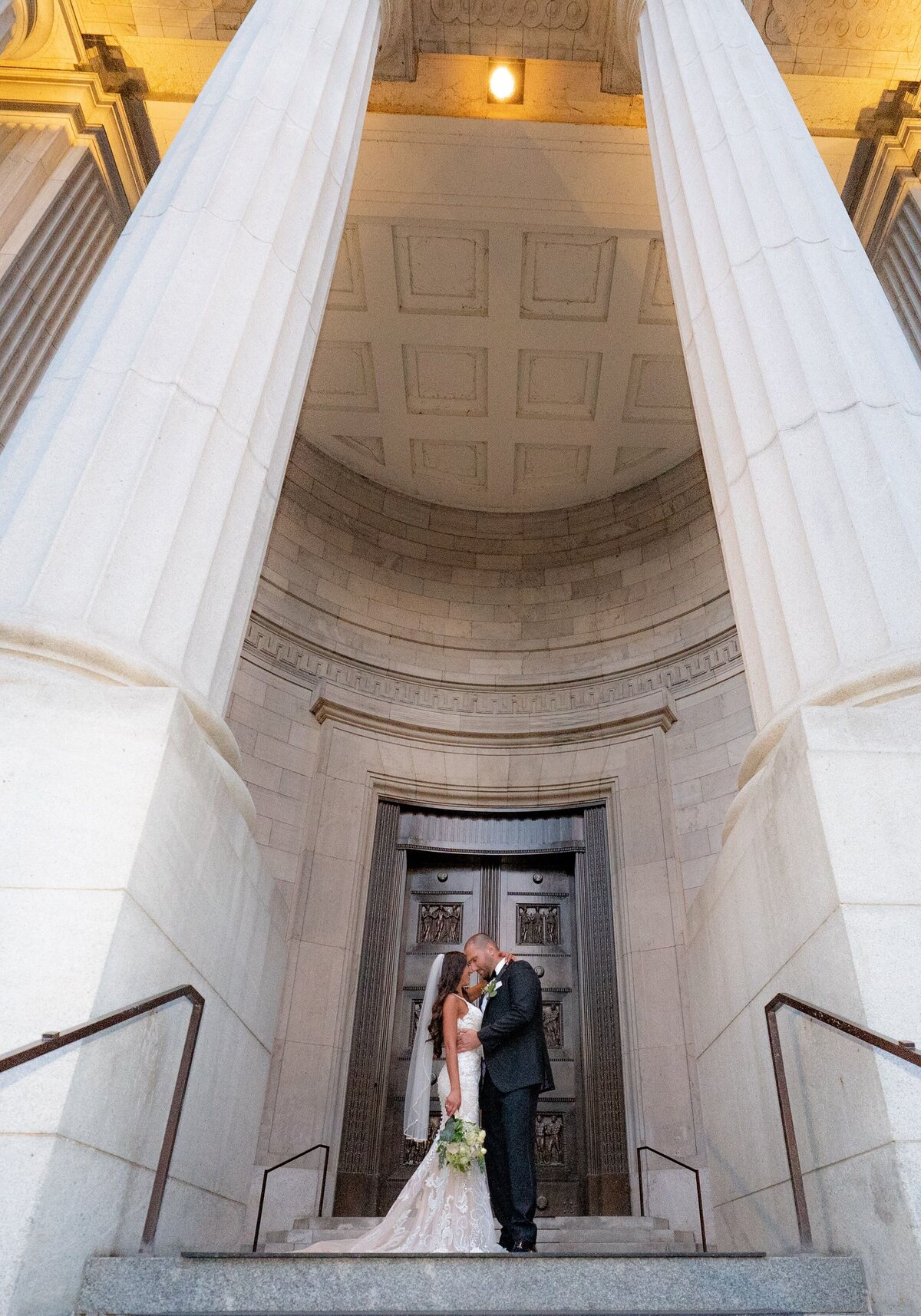 The bride and groom share a romantic kiss in a grand building entranceway adorned with majestic Roman columns. The architectural grandeur of the space frames their intimate moment, enhancing the elegance and timelessness of their wedding celebration. Their attire and the classic setting create a sophisticated and memorable atmosphere.