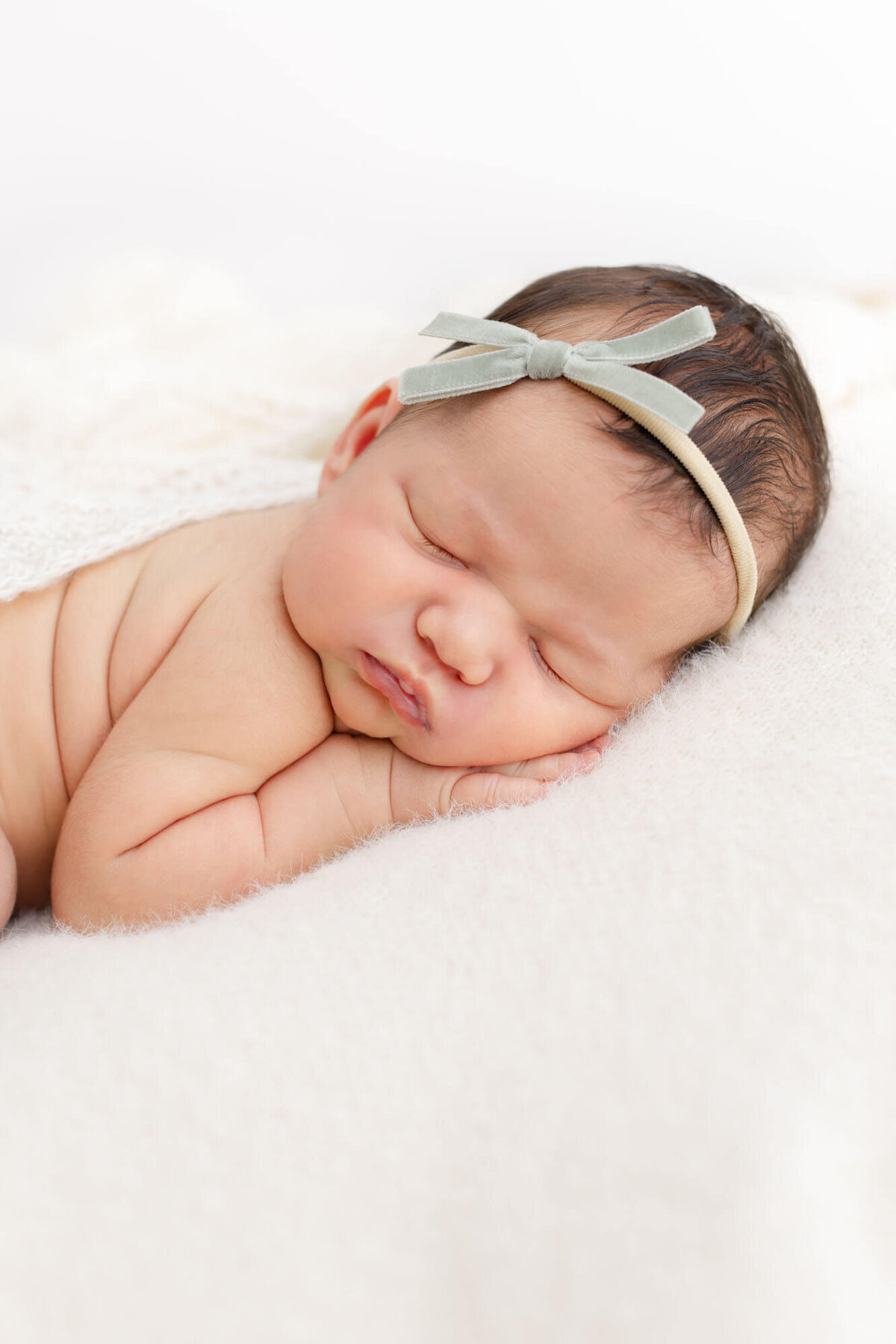 Newborn baby laying on belly with hand up by face wearing a green velvet bow.