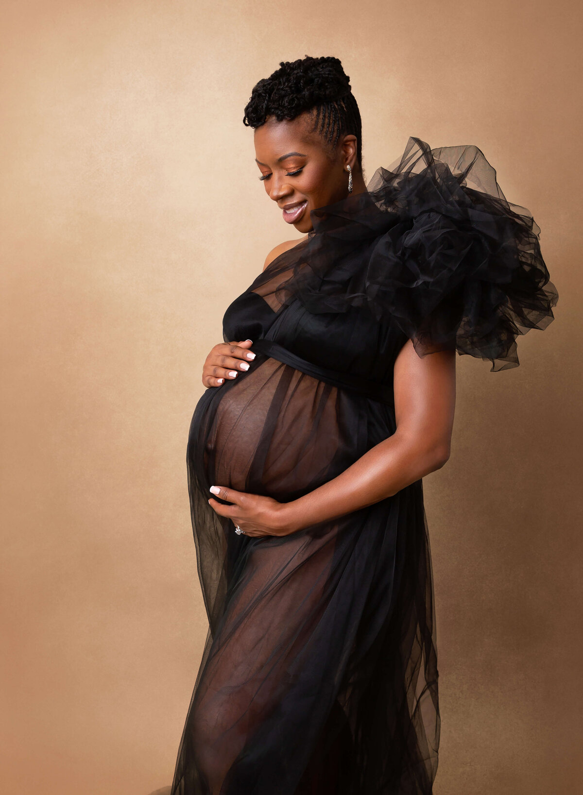 A smiling mother-to-be gazes down at her bump while holding it in a studio in a black tulle gown after a pregnancy photoshoot in New York
