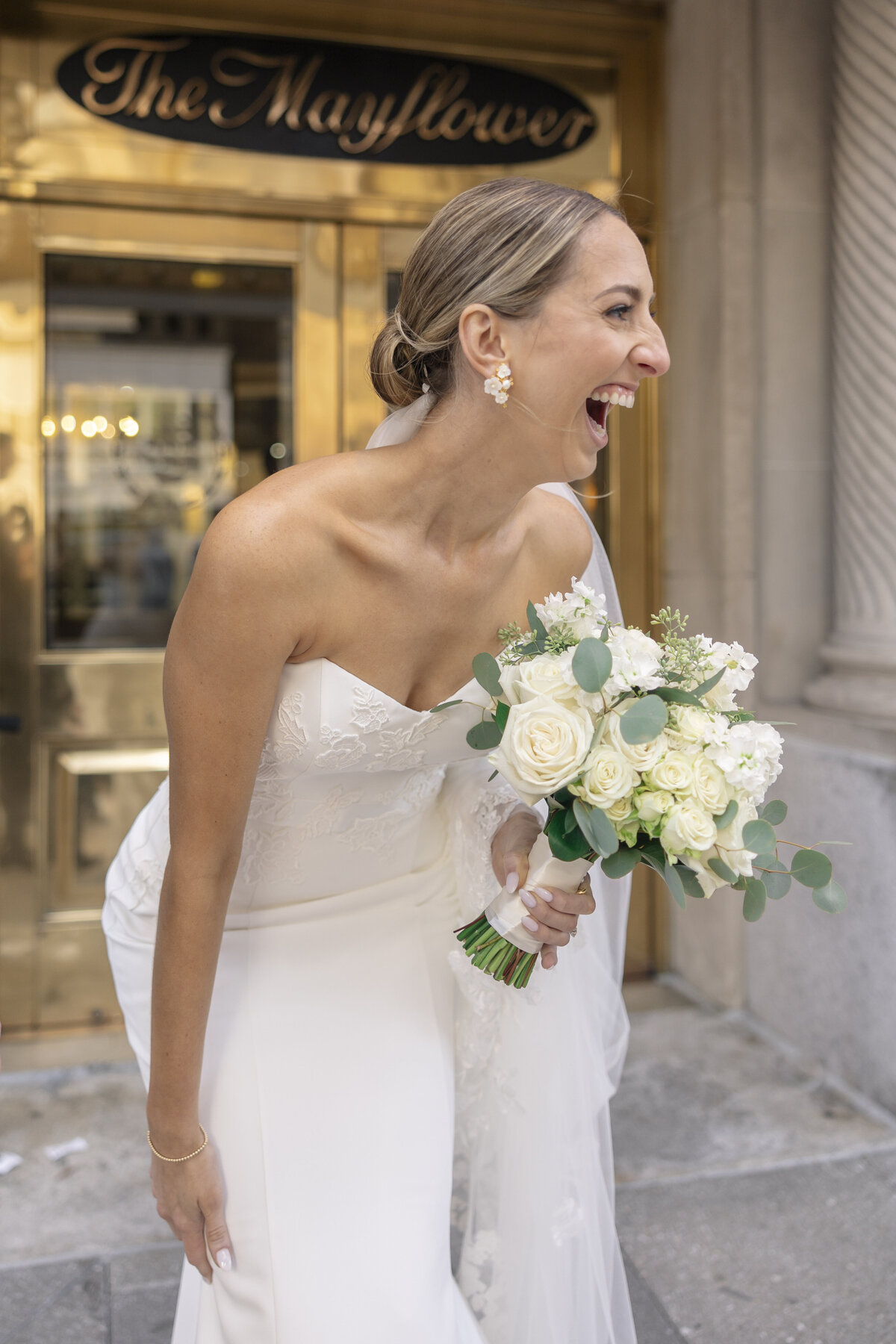 A joyful bride in a white strapless gown laughs heartily while holding a bouquet of white roses and greenery. She stands outside a building with The Mayflower sign above the entrance. Her hair is elegantly styled back.
