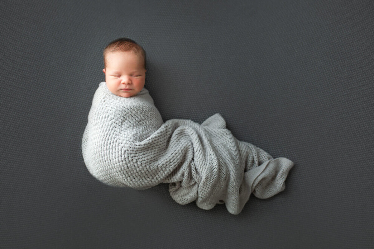 A newborn baby boy sleeps while swaddled under a gray blanket