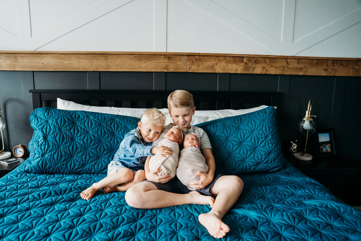 Newborn Photographer, an older brother is hugged by a younger brother as he holds onto twin siblings on parent's bed