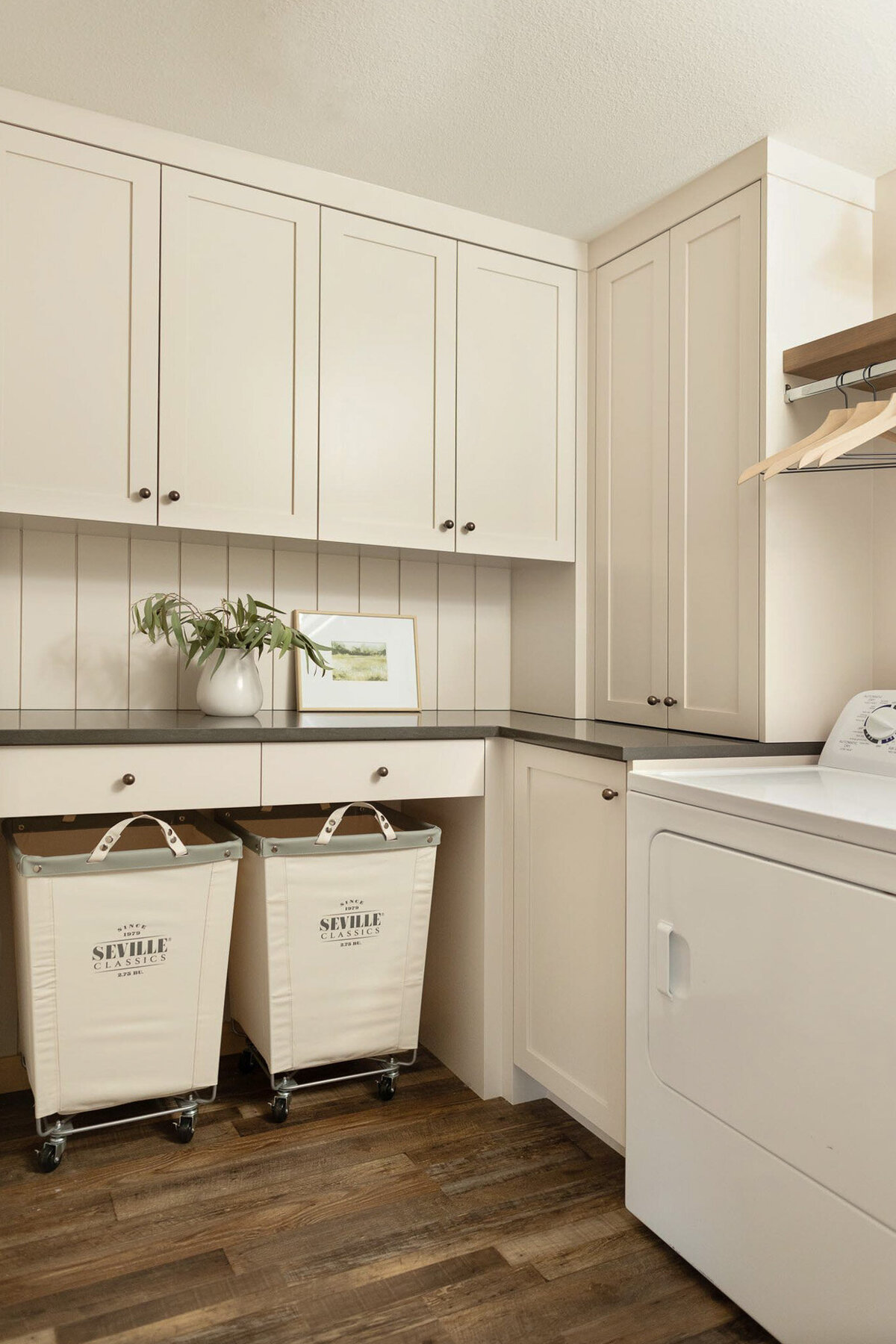 Laundry room with dark brown wood floors. Open shelving area with upholstered laundry hampers on casters. Beige cabinetry and shiplap walls. Beige cabinet with doors with antique bronze knobs. Grey quartz countertops above with open shelving painted beige. Shelves adorned with glass jars of laundry detergent and pods, cleaning spray bottles, towels, jars, and wicker baskets.
