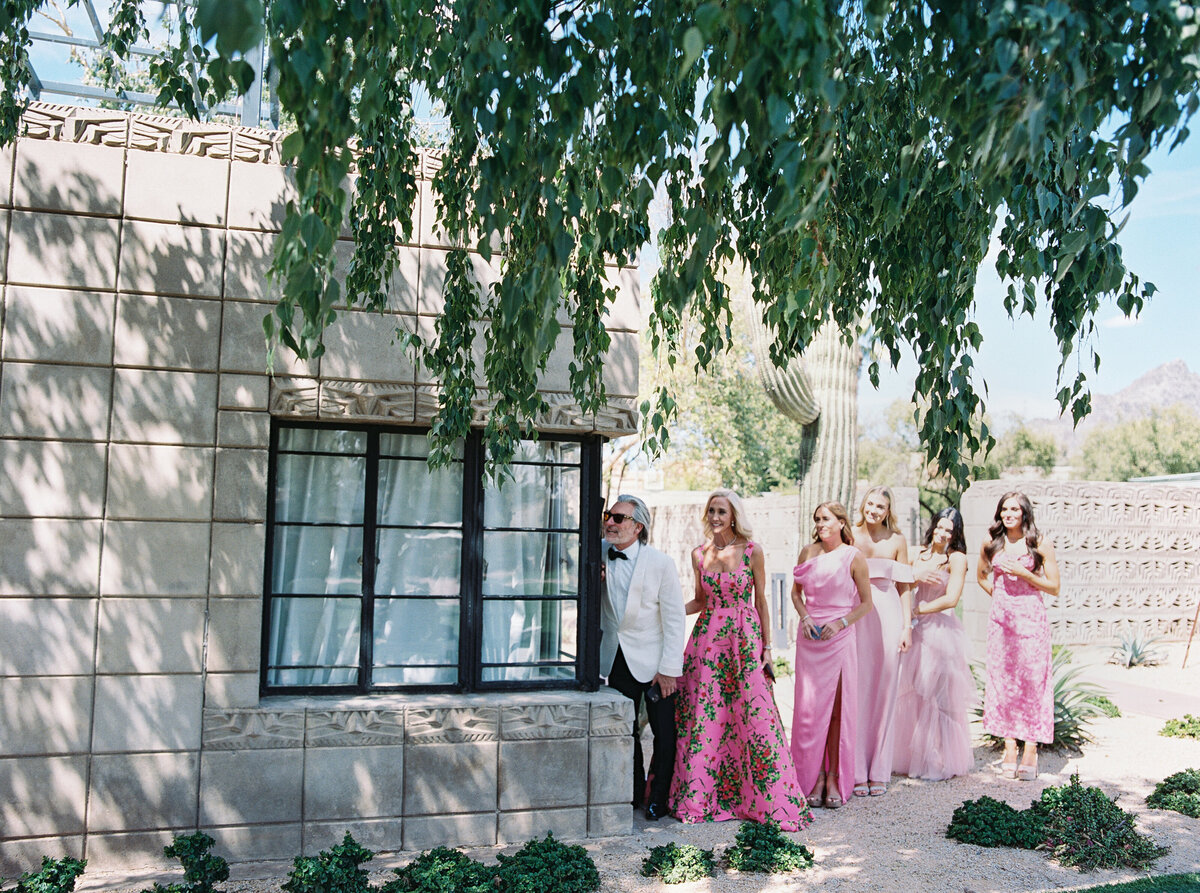 A touching moment as the father of the bride, dressed in a white tuxedo, eagerly peeks through a window for the first look at his daughter on her wedding day.