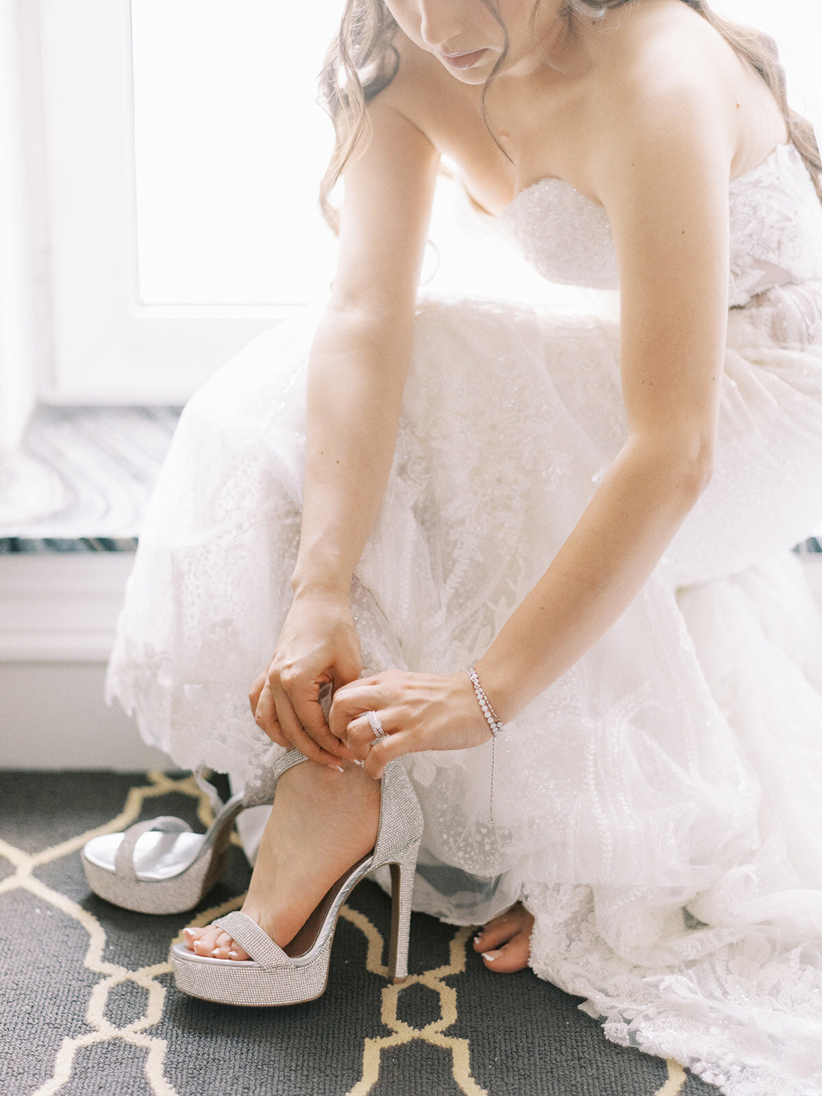 A bride in a strapless wedding gown adjusts her high-heeled, open-toe shoe while sitting on a patterned floor at her classic Calgary wedding.