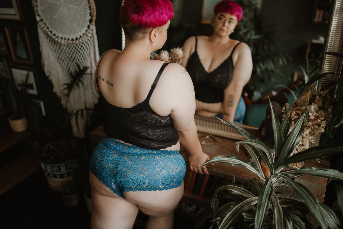 A woman in a black bra and blue underwear looks at herself in a mirror during a boudoir session in Vancouver, BC.