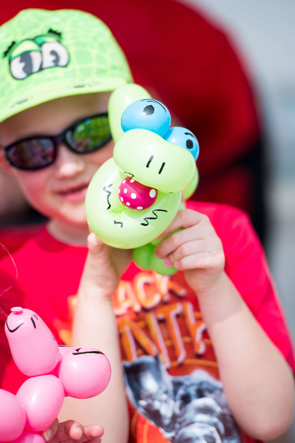 A little boy in a red t-shirt and green cap showing off his balloon animal during a corporate children's event.  Captured by Ottawa Event Photographer JEMMAN Photography COMMERCIAL