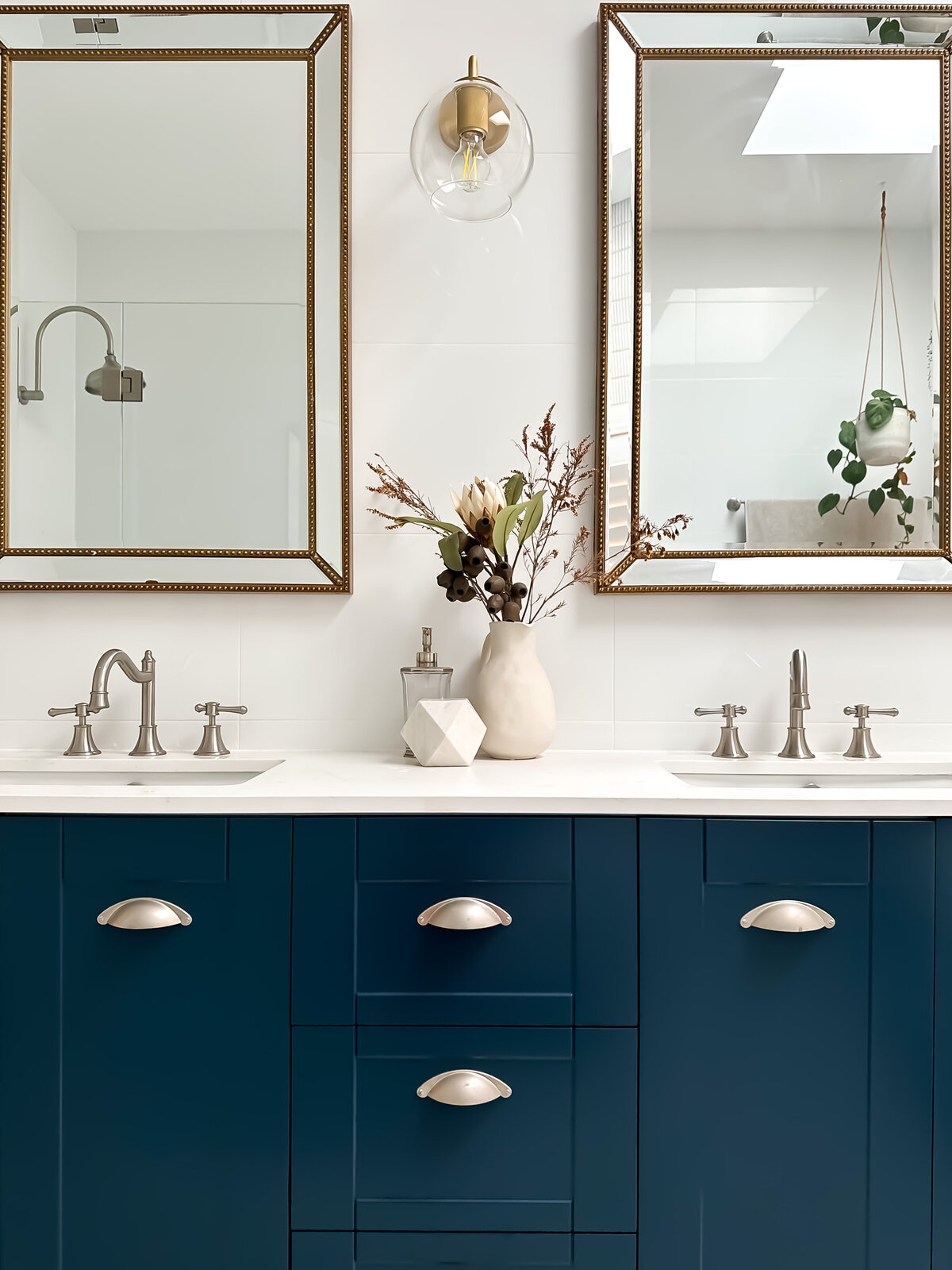 Elegant bathroom designed by Newcastle Kitchen and Bathroom Designer featuring double sinks, blue cabinets, gold-framed mirrors, and a decorative vase between faucets.