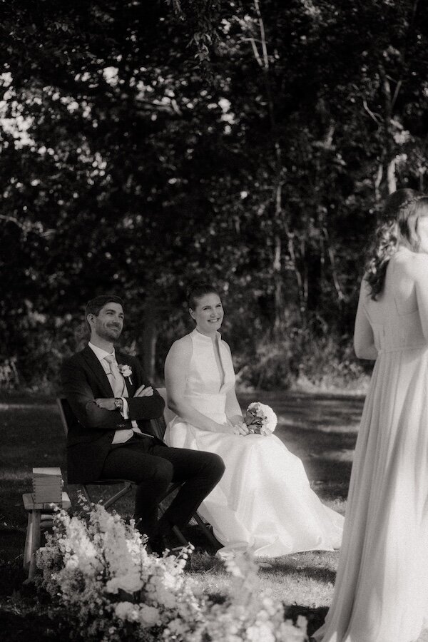 Clarisse et Corentin, assis côte à côte sous les arbres pendant un discours de la cérémonie. Photo noir te blanc pat Kamescop, photographe de mariage en Bretagne.
