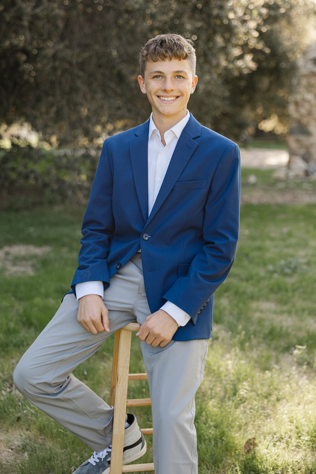 Teen boy sitting on a wooden stool outdoors, wearing a blue blazer and light gray pants. Photographed by Yucaipa family photographer Kaitlyn Dawn Photography.