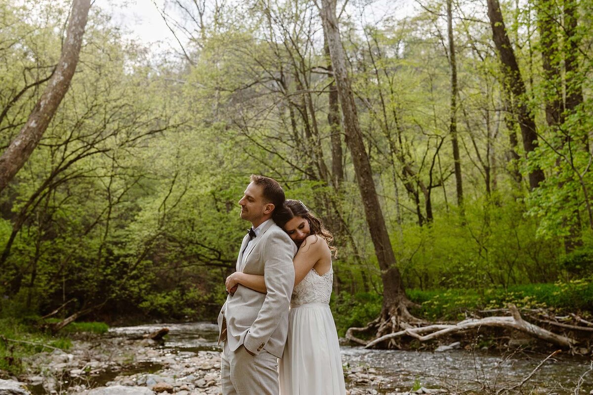 A bride standing behind her groom, and emotional snuggles with him. They are standing on a river bend, and there is a calmness to this photo