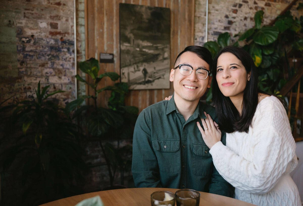 when to take engagement photos in the fall with man and woman in a coffee shop with man sitting on a barstool at a table with woman holding his shoulders and leaning into the man and resting her face to his captured by Virginia wedding photographer