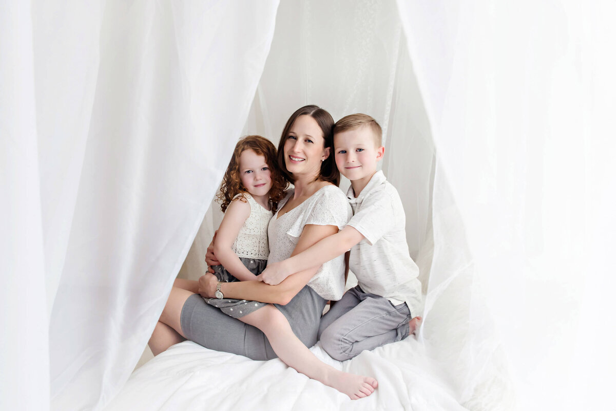 Mom with her son and daughter sitting on a white bed for their mom and me  photography session in Burlington, ON