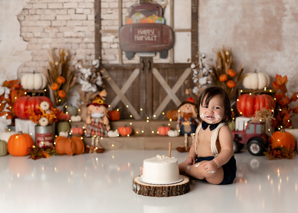 Fall harvest themed cake smash in West Palm Beach, FL newborn photography studio. Baby boy is sitting behind white cake smiling at the camera with cake on his face. In the background, there are rustic barn doors, pumpkins, and tractors.