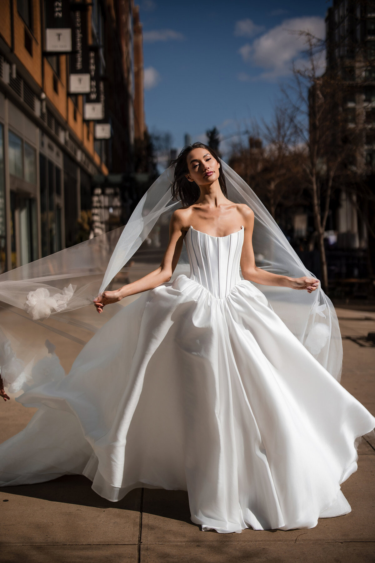 Bride in a strapless structured wedding gown with a flowing veil poses outdoors in bright sunlight on a New York City sidewalk, with modern buildings and bare trees in the background.