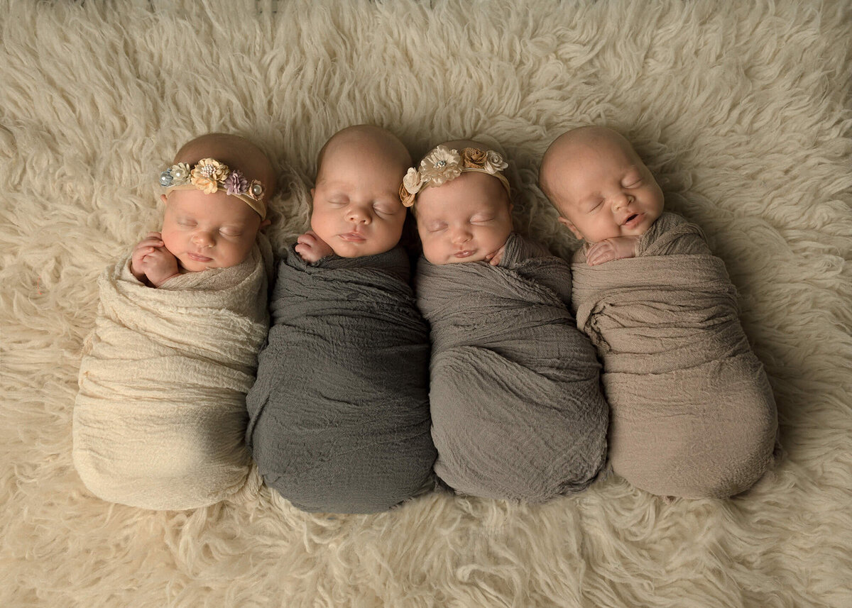 Set of quadruplet newborns all wrapped in neutral wraps laying next to each other on a creme colored rug.