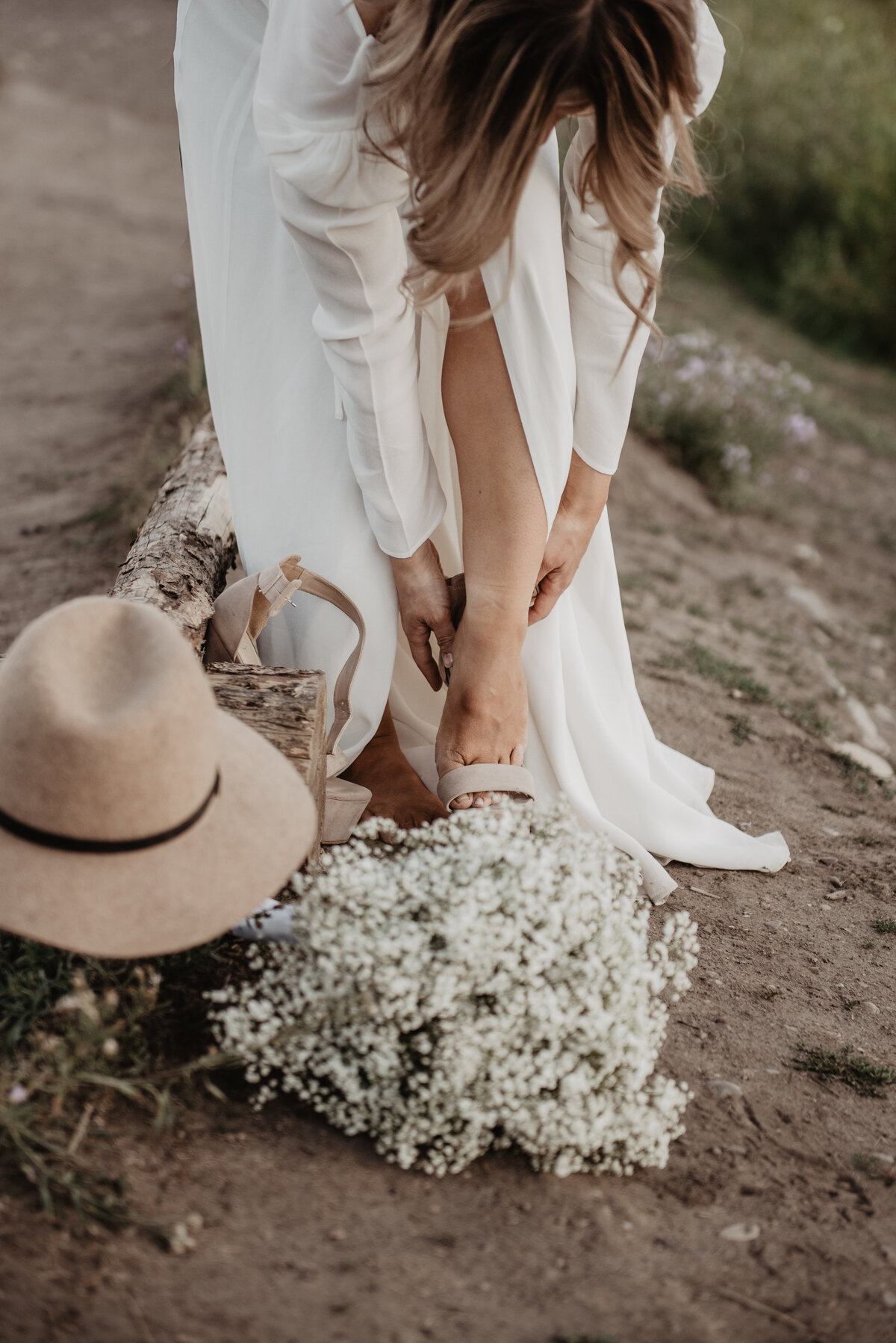 jackson wyoming photographer captures Bride putting on her wedding shoes in the Tetons with her hat and white floral bouquet sitting next to her
