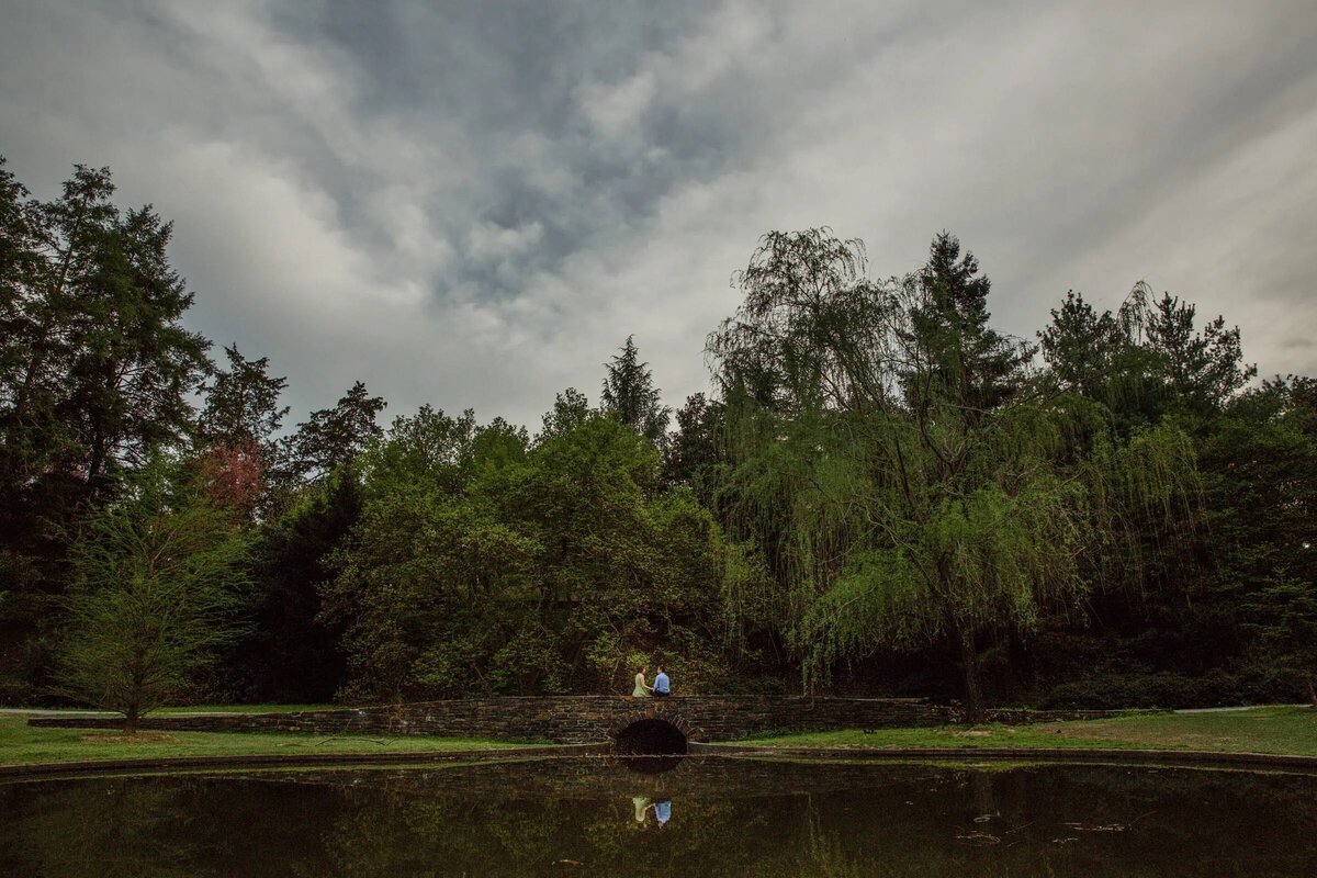 Wide-angle shot of a couple standing beside a reflective pond in a tranquil, expansive garden under an overcast sky