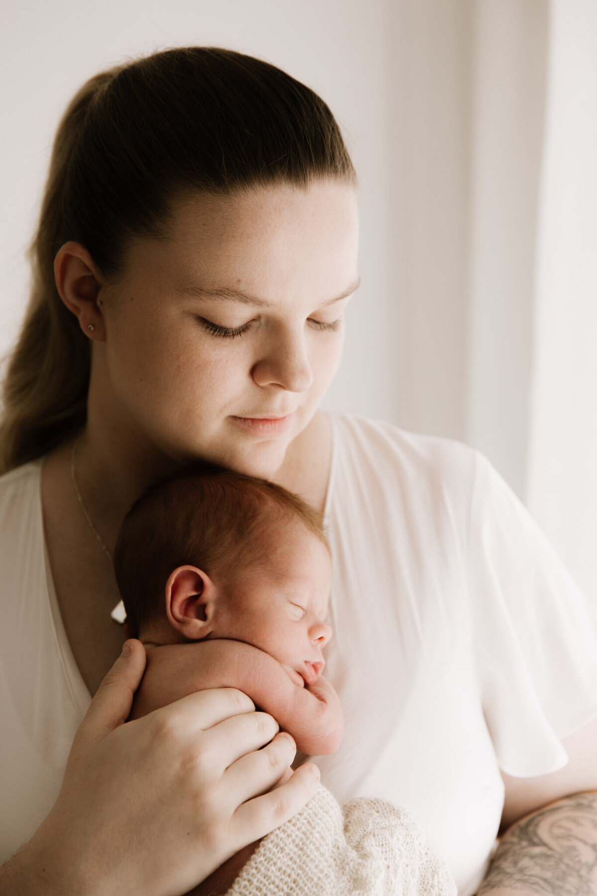 A serene moment of a mother cradling her sleeping newborn close to her chest, captured in soft, natural-style light. The mother wears a white dress and gazes down lovingly, her expression calm and protective. The baby is wrapped in a knitted blanket, resting peacefully. The backdrop features sheer curtains and a white wall, enhancing the warm and intimate feel of the image.