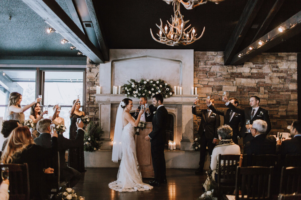 A warm and intimate wedding reception at The Lake House in Calgary, viewed from outside through large glass windows. Guests are seated at long, candlelit tables, engaged in conversation and enjoying the elegant atmosphere. The rustic charm of the venue is highlighted by stone walls, a grand fireplace adorned with greenery and candles, and antler chandeliers casting a soft glow. The dim lighting and reflections in the glass add depth to the scene, creating a cozy and romantic ambiance for the celebration.