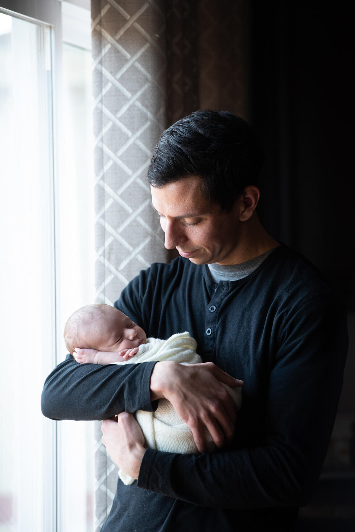 A father stands in a nursery window cradling his sleeping newborn baby in his arms