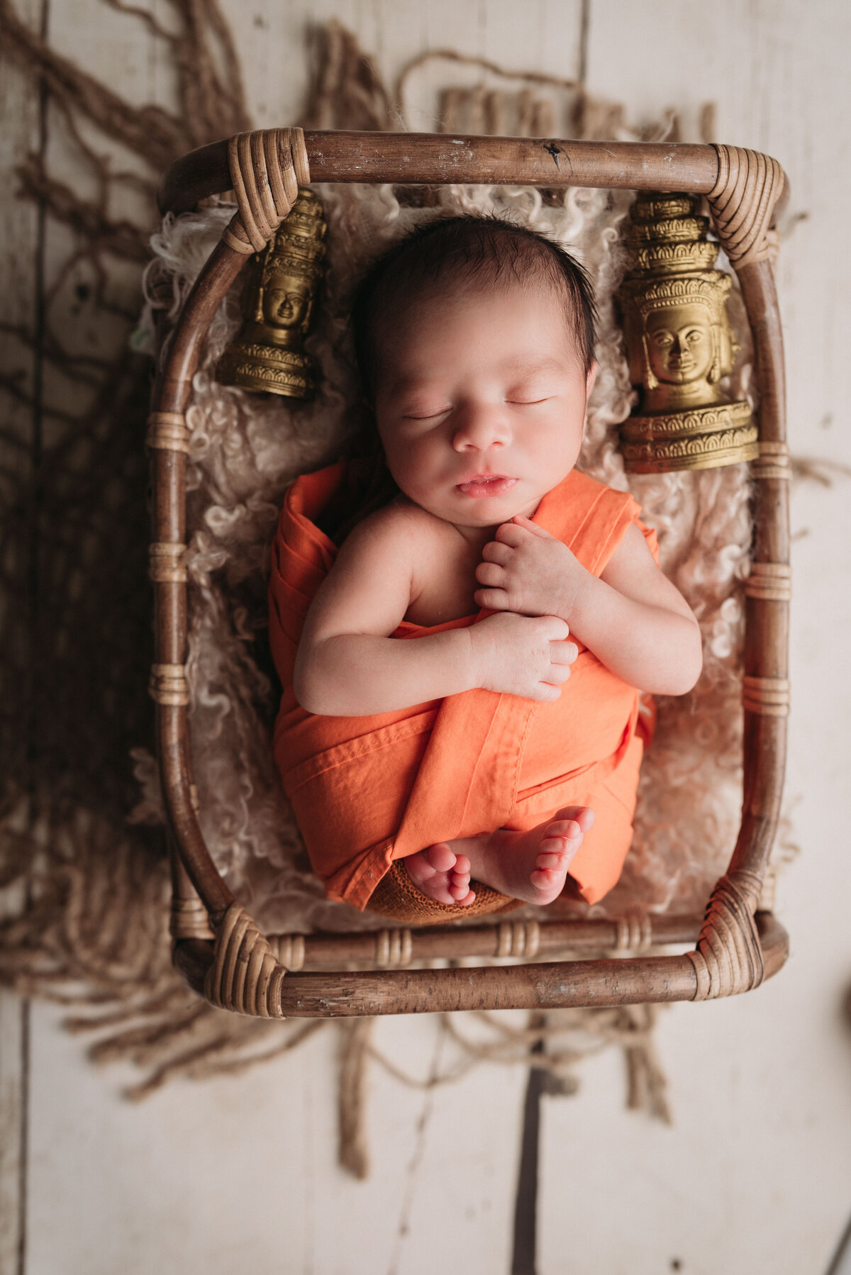 1 week old baby boy in orange wrap laying in brown basket with fur underneath him on white floor backdrop