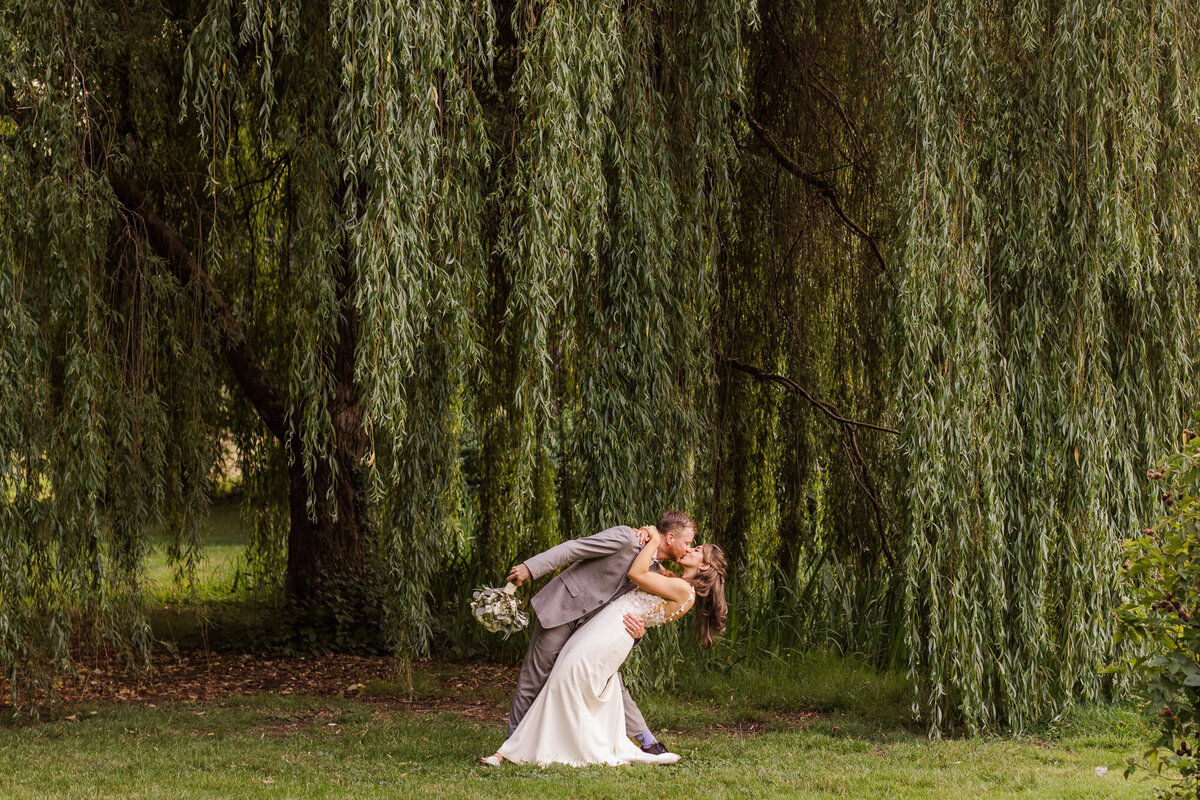 Bride-and-groom-kiss-in-front-of-willow-tree-walk-at-gardens-at-Froggsong-venue-at-Vashon-Island-WA-near-Seattle-photo-by-Joanna-Monger-Photography