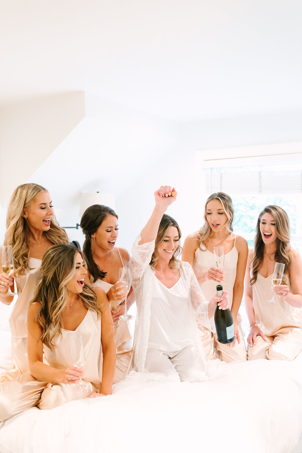 bride with her bridesmaids in soft silk pajamas smiling and opening a bottle of champagne on a hotel bed.